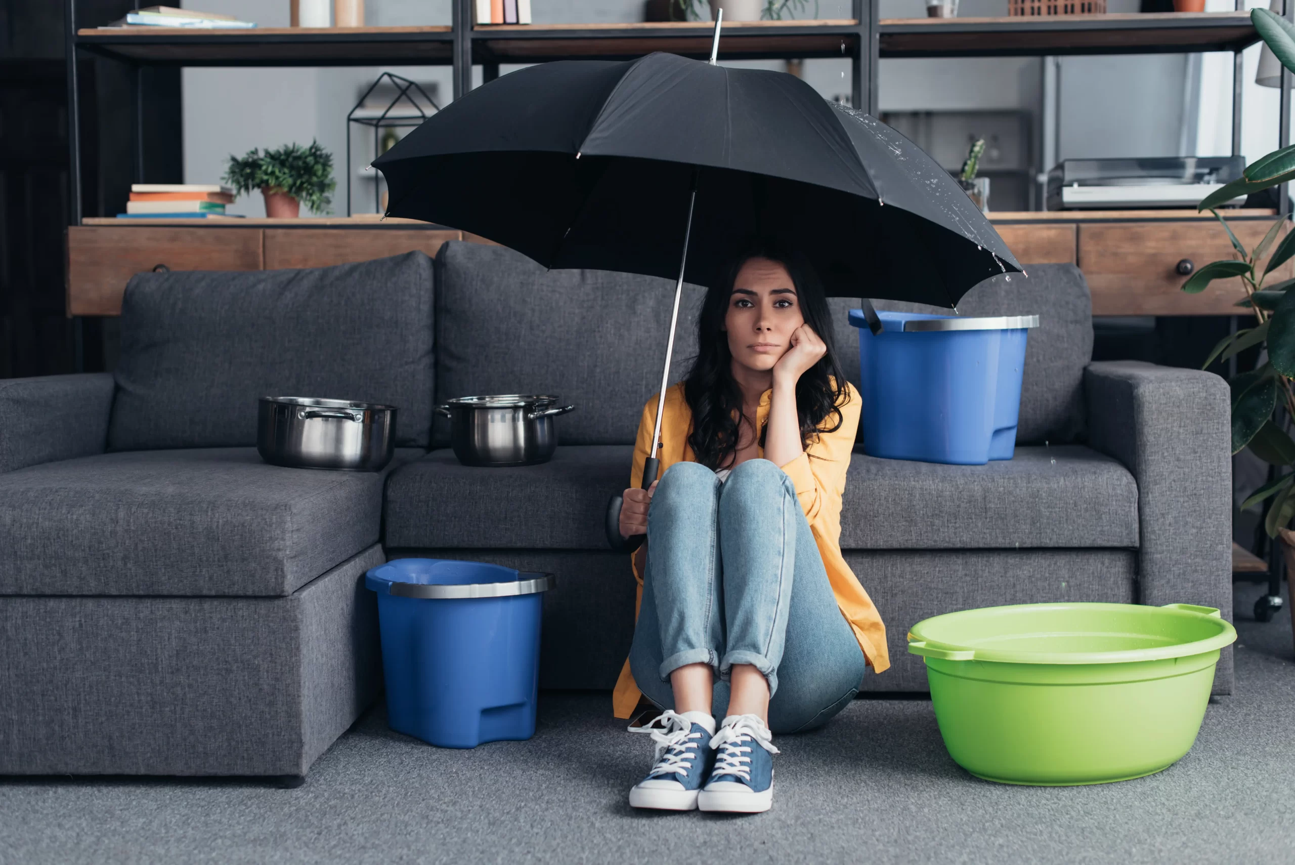 A woman sits on the floor under an umbrella indoors, surrounded by buckets and pots catching water from a ceiling leak. Thankfully, Water Damage Restoration Experts are on their way to transform her soggy situation into a dry and comfortable space again.