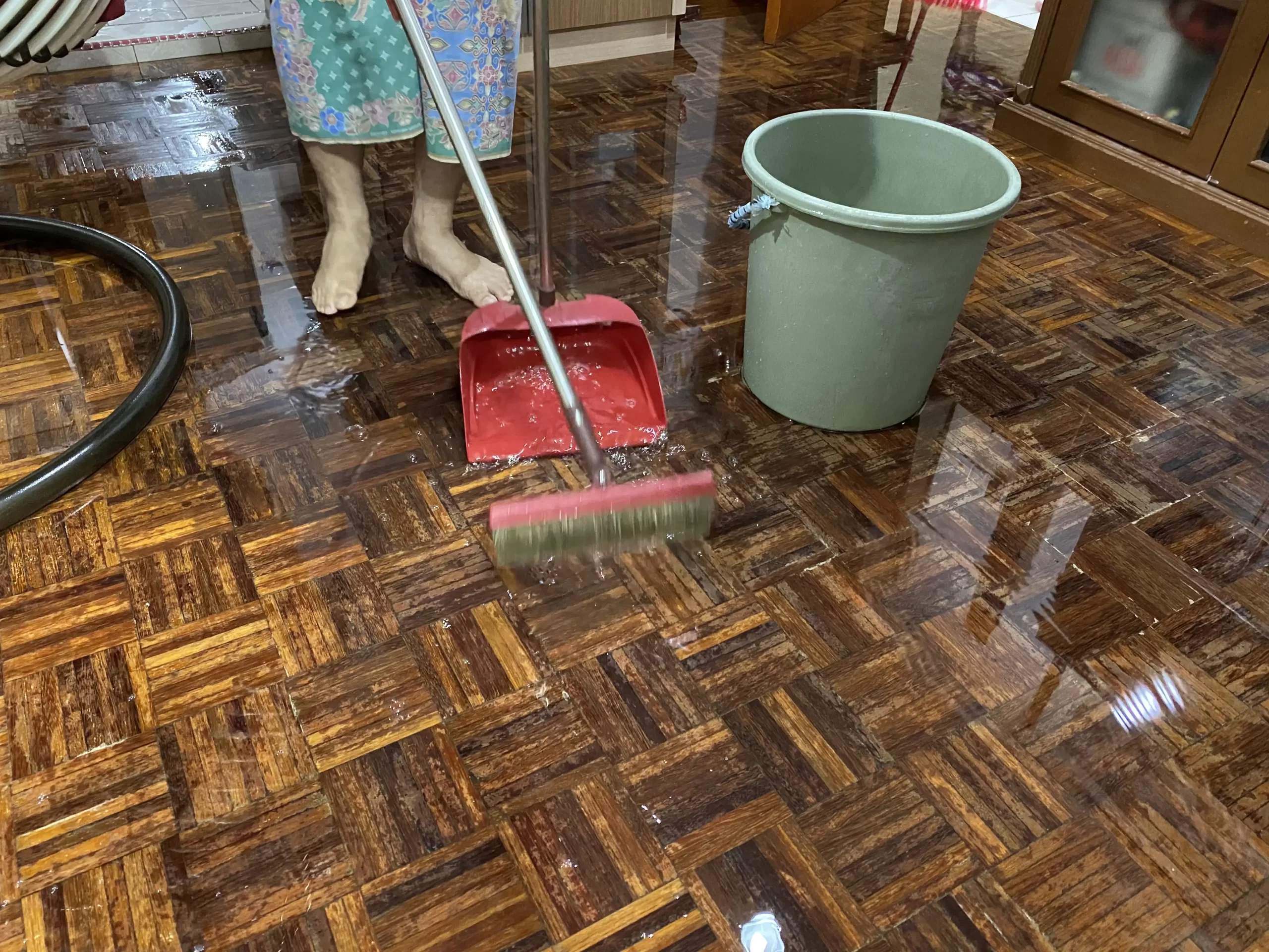A person is sweeping water off a flooded wooden floor with a broom and dustpan, highlighting the urgent need for Water Damage Restoration Experts. Nearby, a green bucket stands ready to assist in the cleanup.