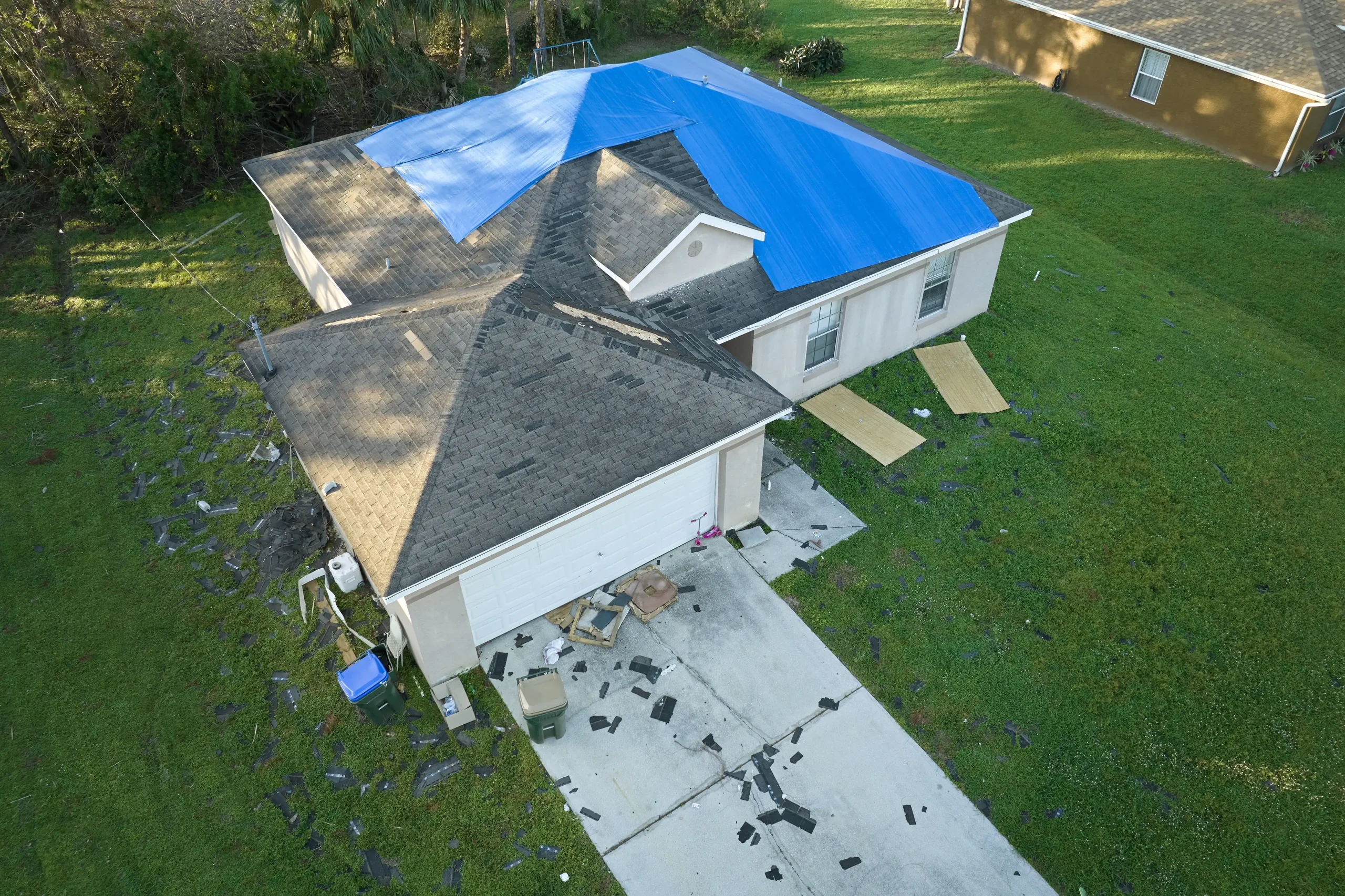 An aerial view of a house in post-storm recovery shows a damaged roof partly covered by a blue tarp, with shingles and debris scattered across the driveway and lawn.