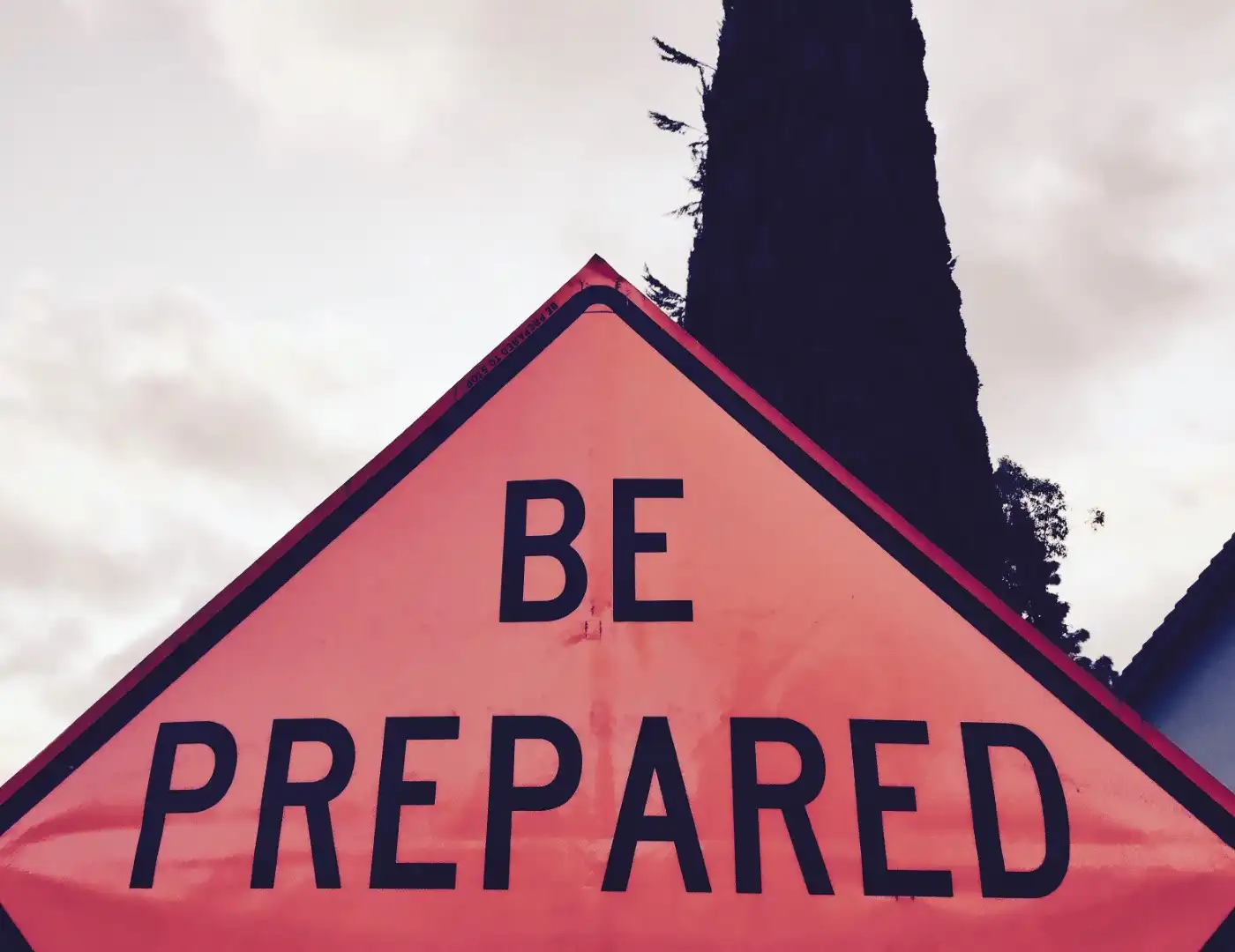 A triangular orange sign with the words "BE PREPARED" stands tall against a cloudy sky and a towering tree, reminding passersby of the essential Storm Response Plan for Commercial Properties.