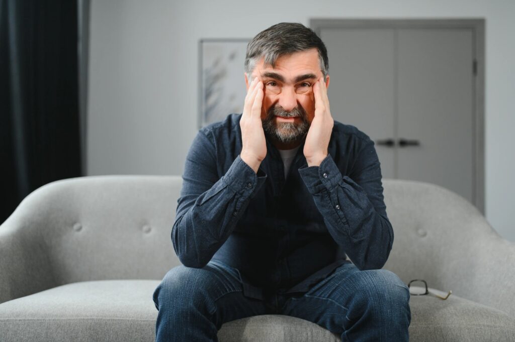 A man sits on a sofa, touching his temples with his hands, looking distressed. Glasses are placed beside him, hinting at a day gone awry. Nearby, a pamphlet for Emergency Restoration Services lies open, suggesting a possible solution to his troubles.