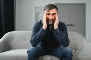 A man sits on a sofa, touching his temples with his hands, looking distressed. Glasses are placed beside him, hinting at a day gone awry. Nearby, a pamphlet for Emergency Restoration Services lies open, suggesting a possible solution to his troubles.