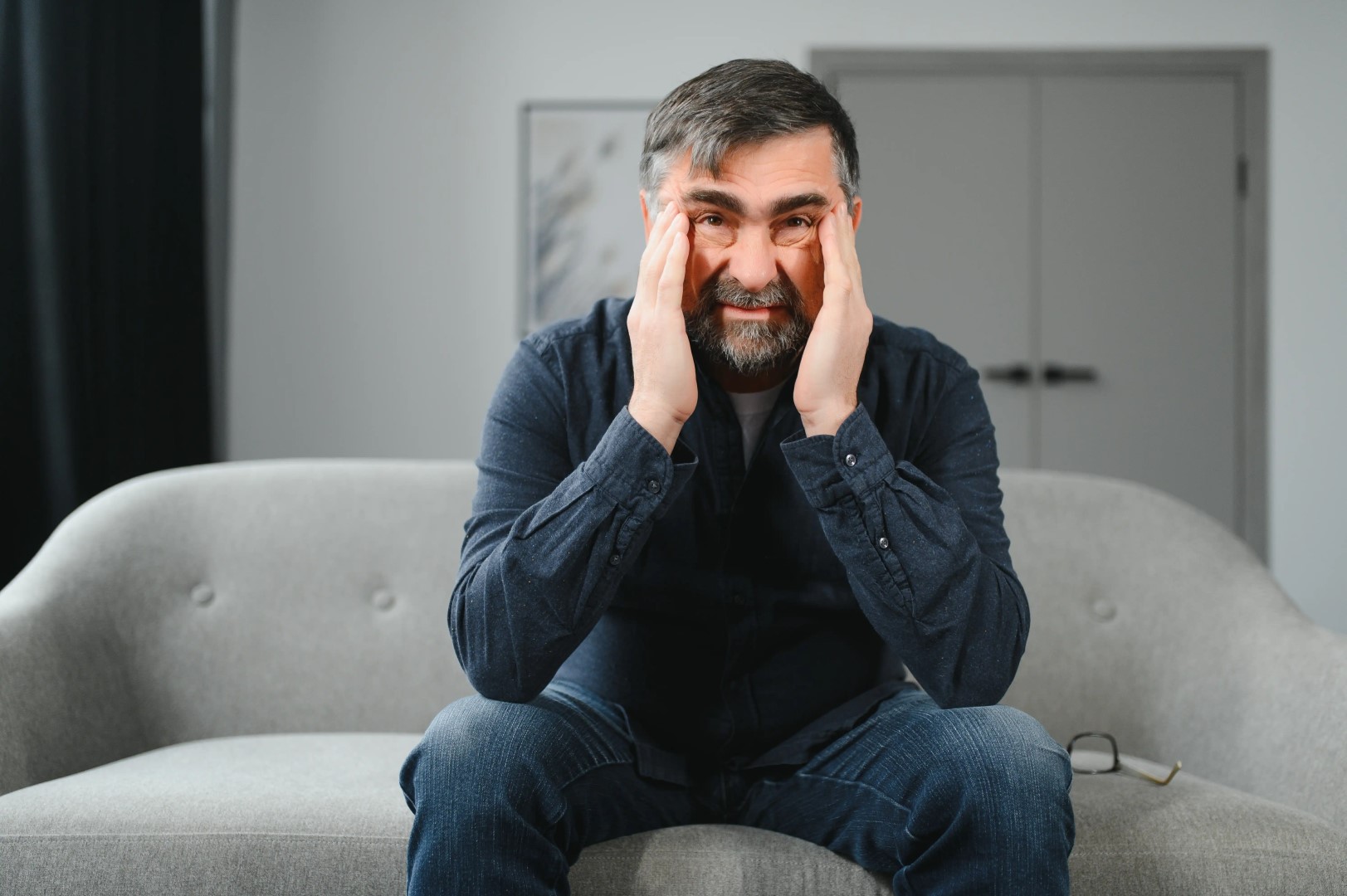 A man sits on a sofa, touching his temples with his hands, looking distressed. Glasses are placed beside him, hinting at a day gone awry. Nearby, a pamphlet for Emergency Restoration Services lies open, suggesting a possible solution to his troubles.