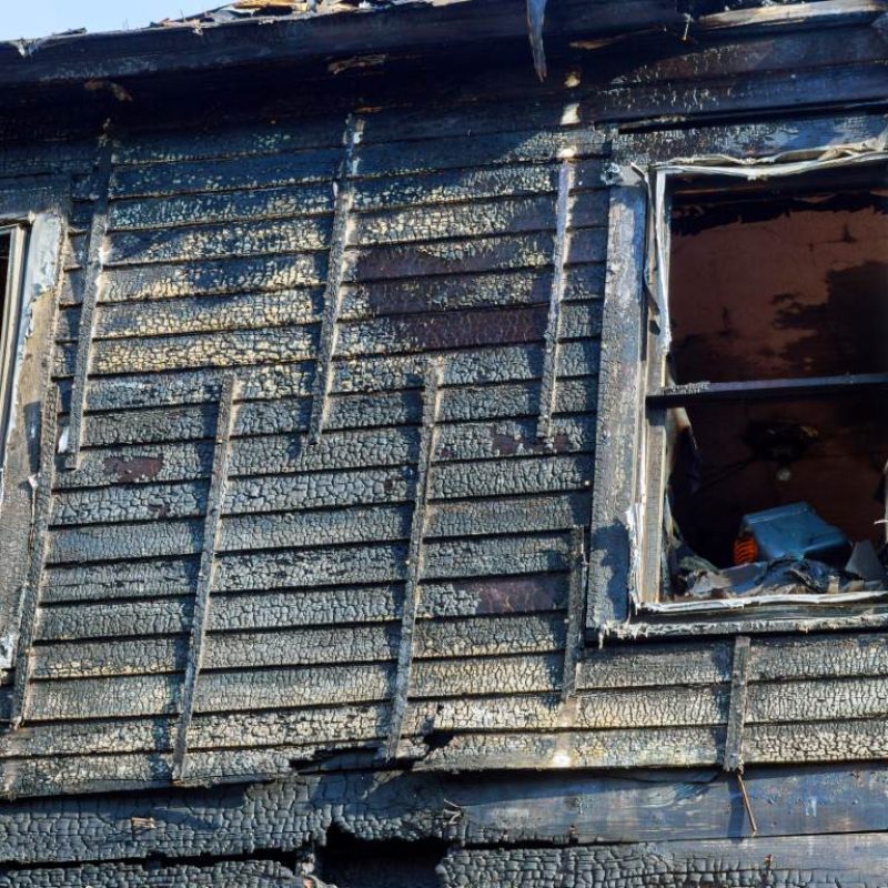 The charred exterior of a burned building with two open windows reveals extensive interior damage, highlighting the urgent need for fire and smoke restoration services.