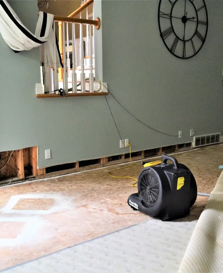 Room under renovation with partially removed flooring, exposed wall sections, and a large fan skillfully placed by American Restoration Contractors. A hammock hangs by the staircase, while a clock is mounted on the wall.